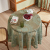 A round table set for a meal with a green floral lace tablecloth, a white teapot, and a bowl of red fruit.