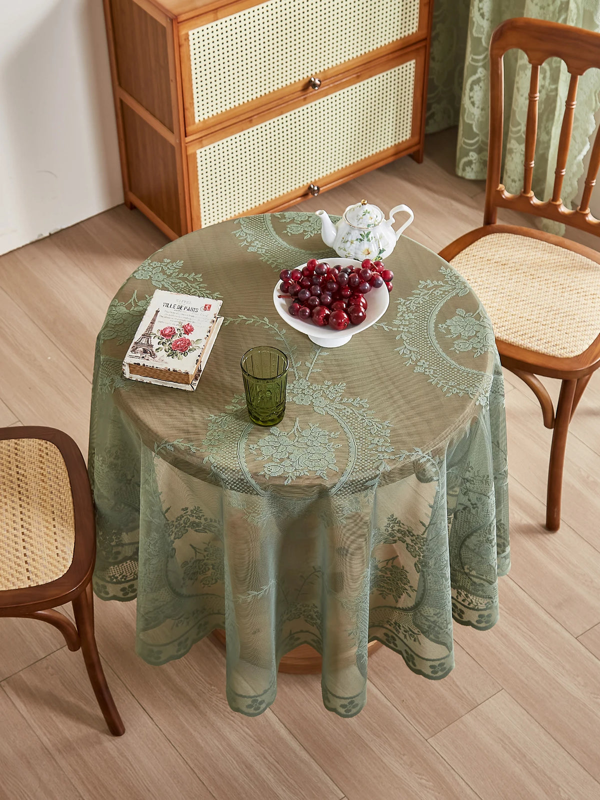 A round table set for a meal with a green floral lace tablecloth, a white teapot, and a bowl of red fruit.