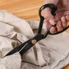 A hand is using heavy-duty carbon steel tailor scissors with an ergonomic black handle to cut fabric.