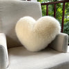 A fluffy, heart-shaped throw pillow made of light cream fox fur resting on a beige sofa.