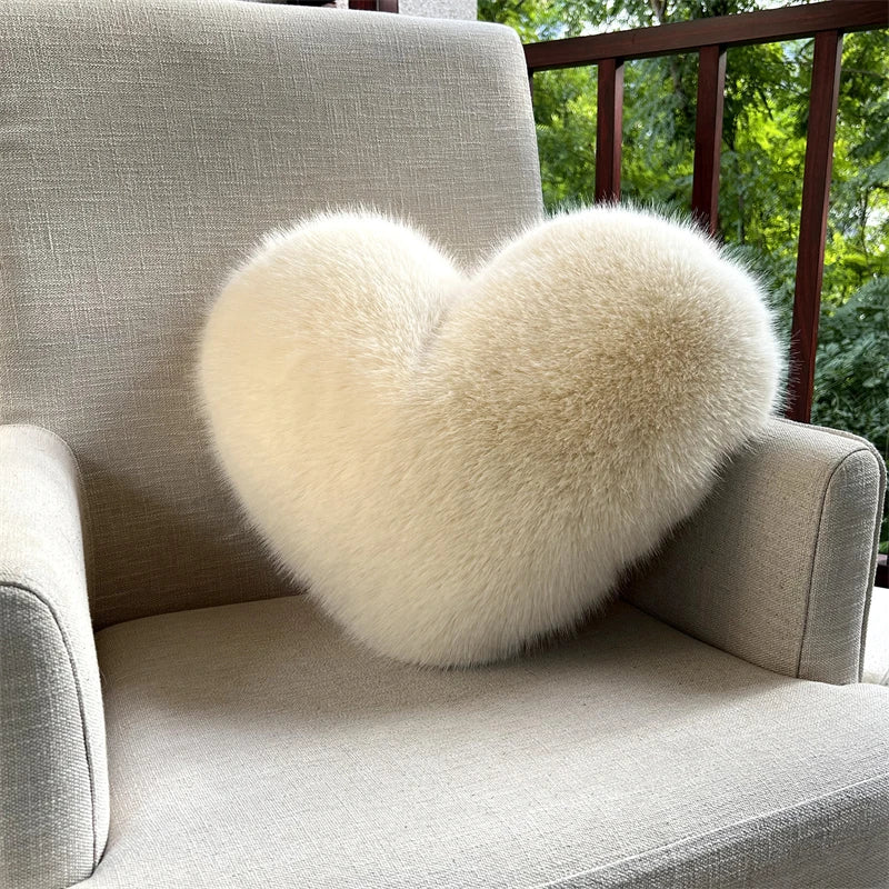A fluffy, heart-shaped throw pillow made of light cream fox fur resting on a beige sofa.