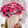 A hand knitted floral rose bouquet made of pink and green yarn is held against a white shirt and yellow garment.