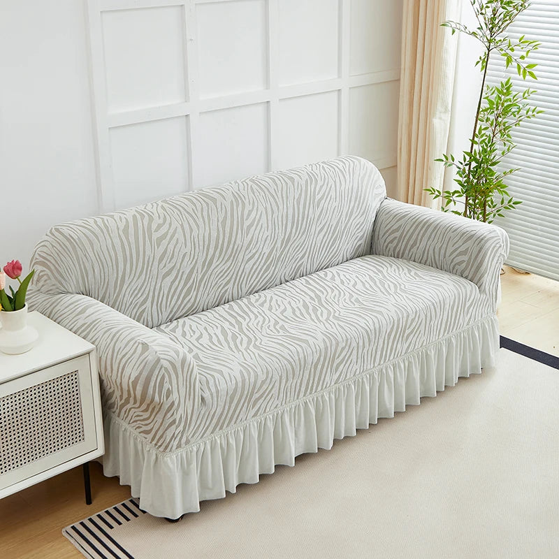 A light gray anti-slip jacquard sofa slipcover with a ruffled hem sits on a white cabinet in a bright room.