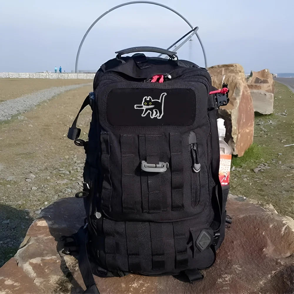 Black tactical backpack with an embroidered cute assassin cat patch resting on a rock.