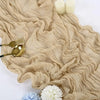 A rustic beige gauze table runner with draped folds, topped with white candles and floral accents.