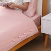 A pink quilt is displayed on a wooden bed frame with a person reading nearby.