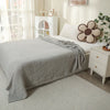 A light gray quilted blanket draped over a bed in a bright, minimalist bedroom setting.