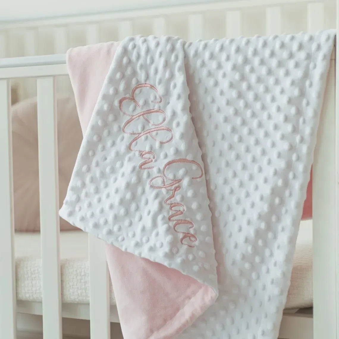 A white and pink personalized baby name swaddle blanket with delicate script embroidery hanging on a wooden crib rail.