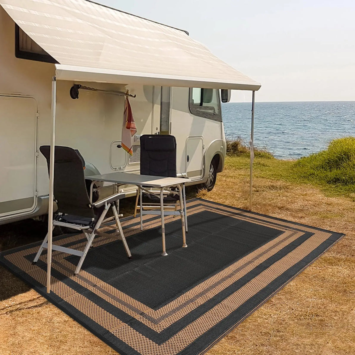 A black outdoor patio rug with striped edges is placed on dry grass in front of a white RV under a canvas awning.