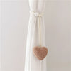 White tieback with a pearl knot and a fuzzy pink heart decoration hanging from a sheer white curtain.