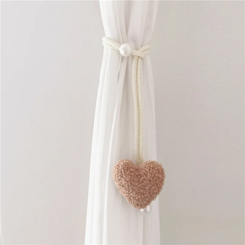 White tieback with a pearl knot and a fuzzy pink heart decoration hanging from a sheer white curtain.