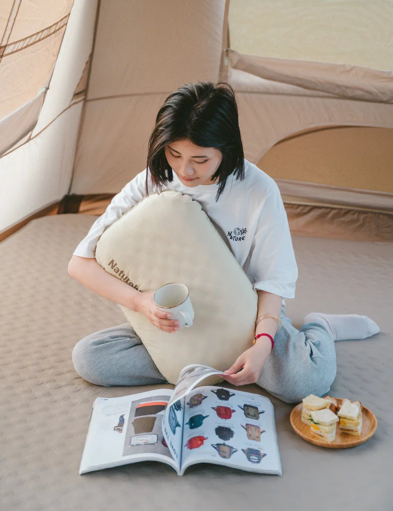 A person is reading a book while resting their head on a beige inflatable camping pillow in an outdoor tent setting.