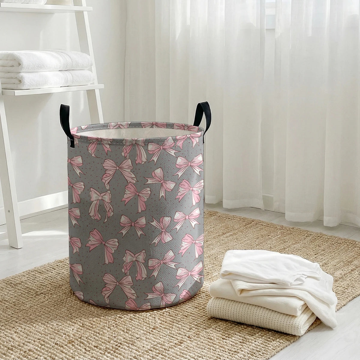A gray laundry basket with a pink bow pattern sits on a woven rug next to sheer white curtains.