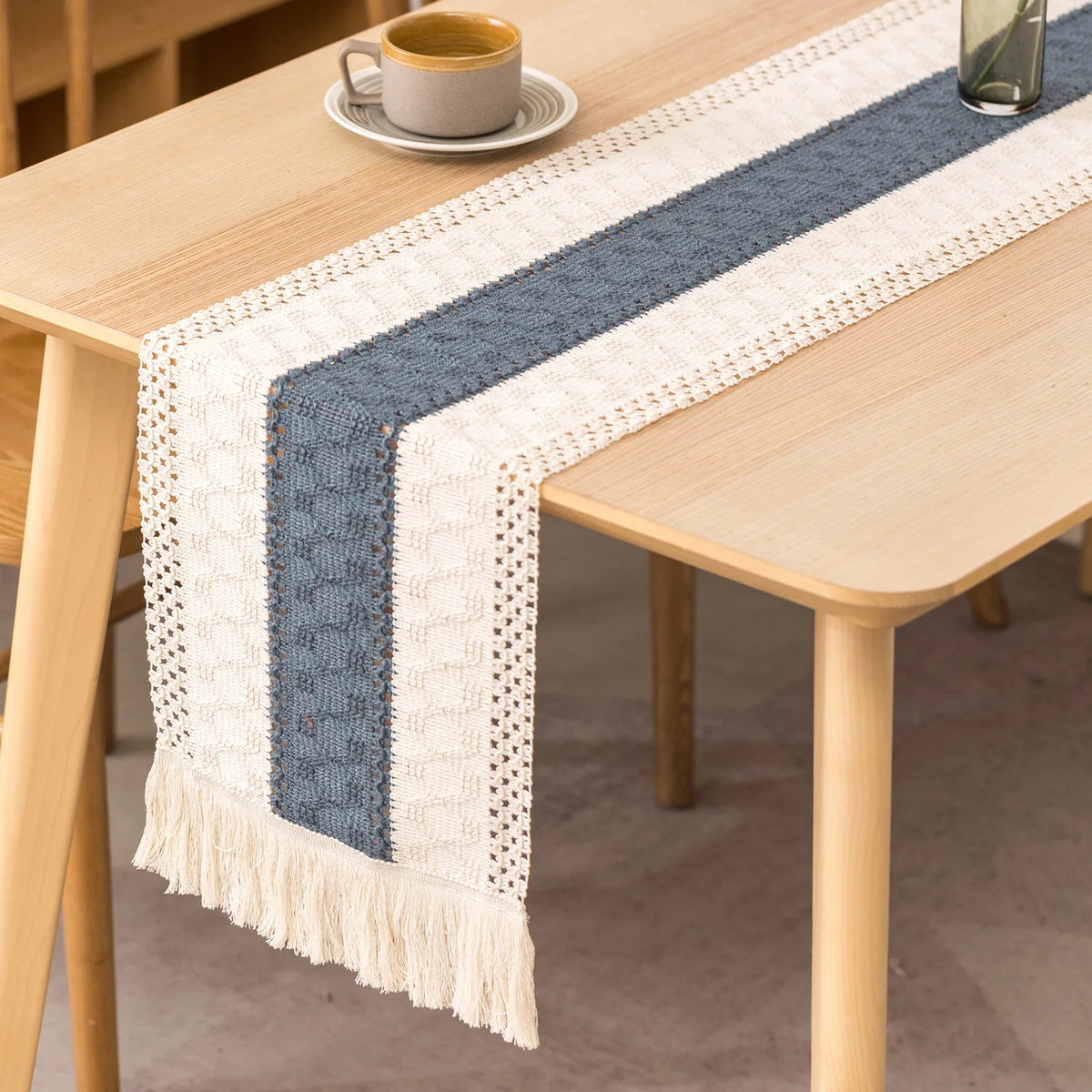 A boho linen cotton table runner with blue and white stripes and tassels placed on a light wood table.