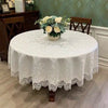 A white embroidered lace tablecloth with an elegant scalloped edge is displayed on a round table surrounded by floral arrange
