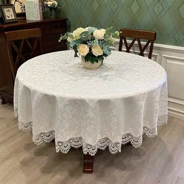 A white embroidered lace tablecloth with an elegant scalloped edge is displayed on a round table surrounded by floral arrange