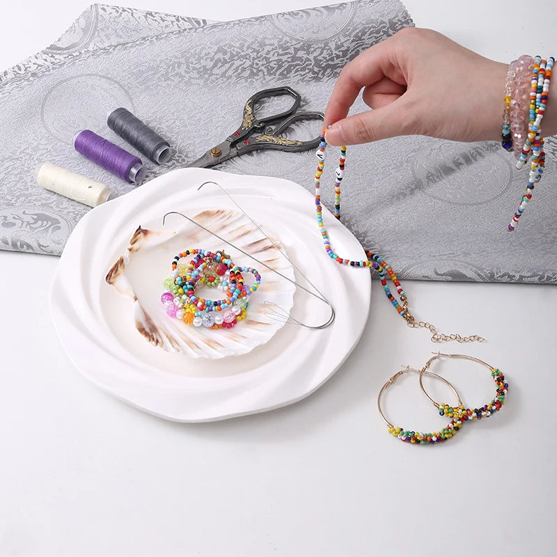 A hand is using precision beading needles to thread colorful beads onto jewelry strings on a white plate.