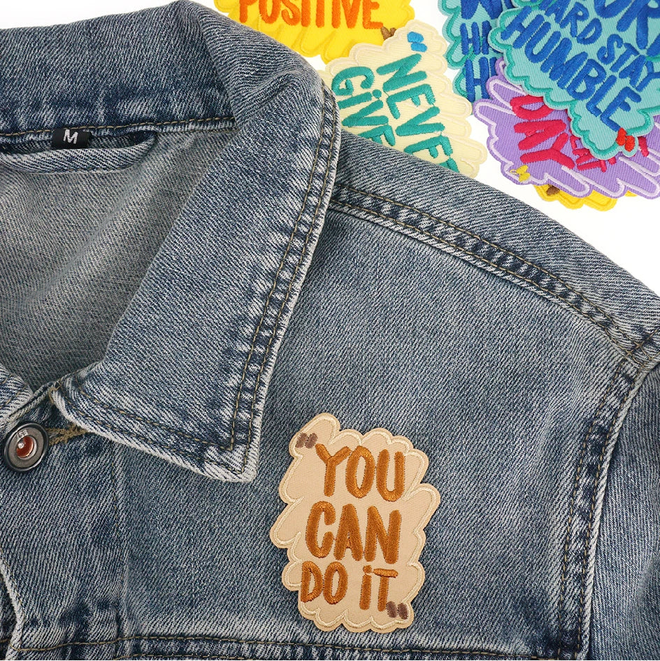 A close-up of a denim jacket featuring several colorful iron-on embroidered text patches.