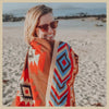 A woman smiling while wearing a patterned knit blanket outdoors on a sandy beach.