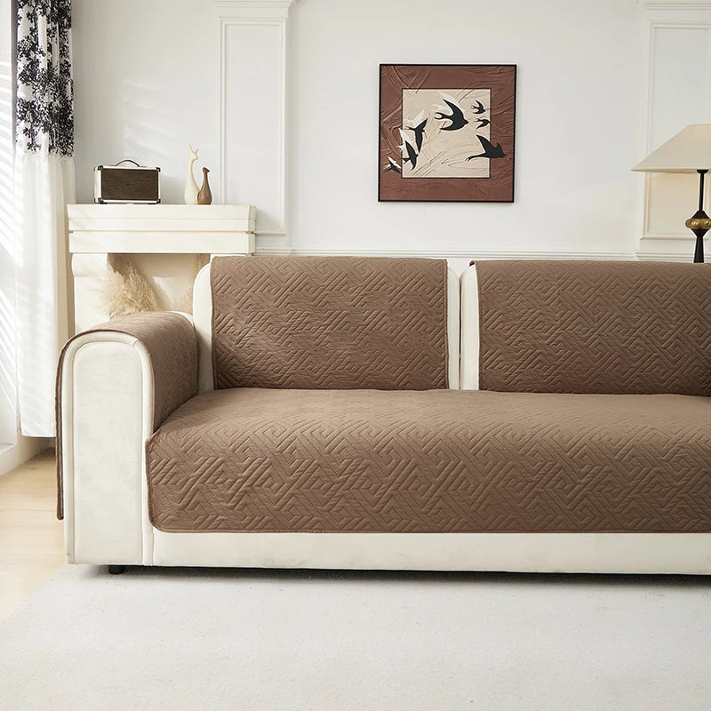 A beige geometric quilted sofa cushion cover on a white frame sits on a light rug in a bright room.