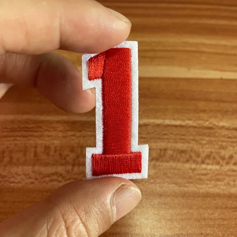 A hand holds a red embroidered number one patch with white stitching on a wooden surface.