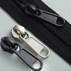 A close-up of a black zipper with silver and black replacement sliders and a separate black slider tool.