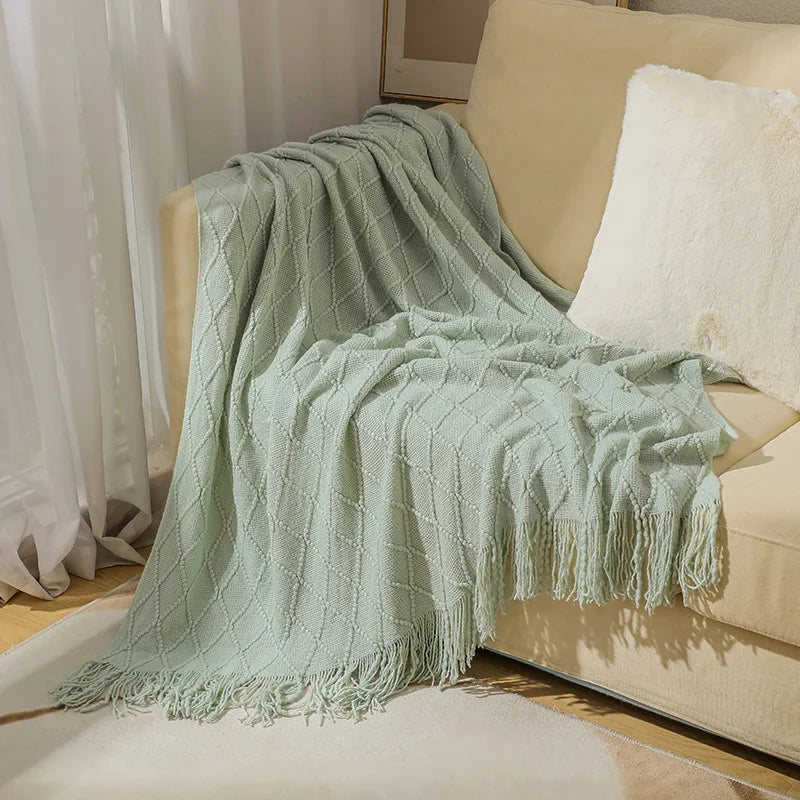 A light sage green knitted tassel throw blanket draped over a beige sofa.