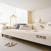 A beige plush sofa cover with a quilted texture is displayed on a bed, next to a modern lamp and sheer curtains.