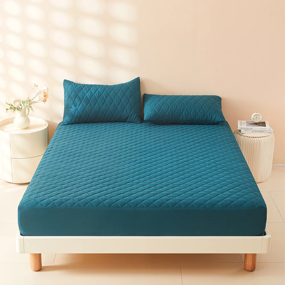 A teal quilted mattress protector cover lying on a white bed frame with pillows and a small vase nearby.