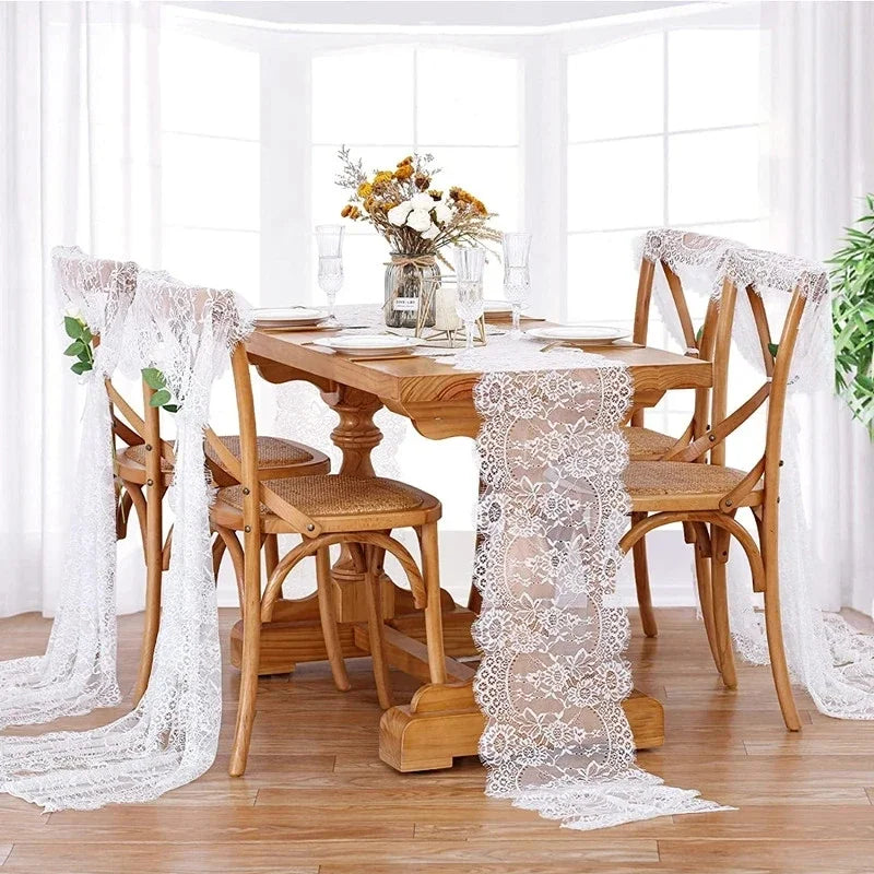 A wooden dining table set with a white embroidered floral lace table runner and sheer white fabric on the chairs.