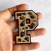 A hand holds a black iron-on patch shaped like the letter P with a leopard print design.