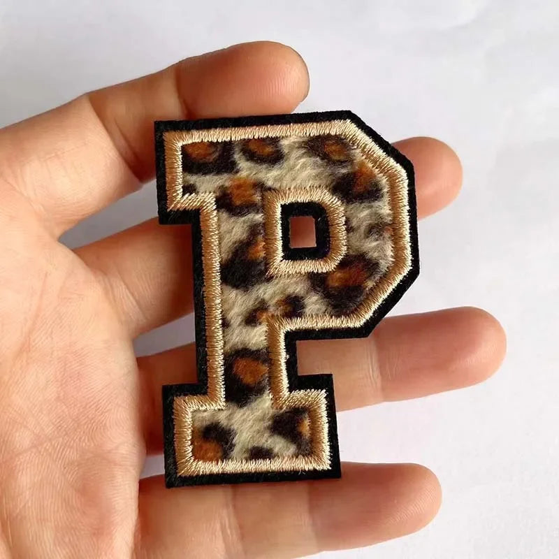 A hand holds a black iron-on patch shaped like the letter P with a leopard print design.