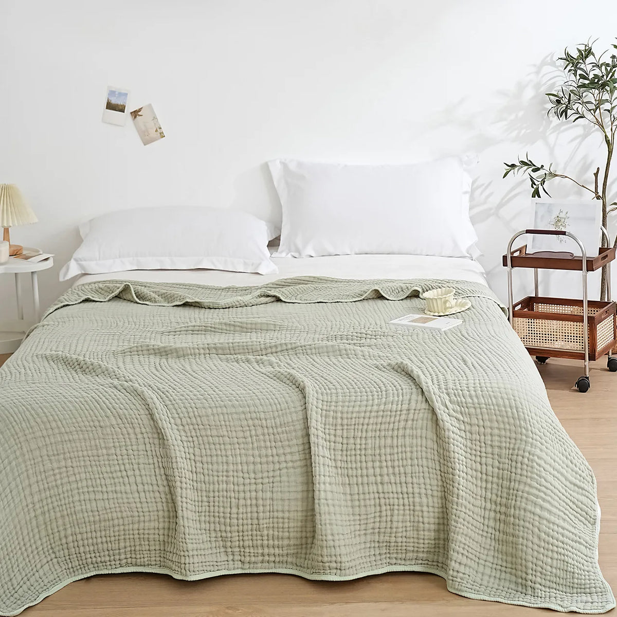A light sage green muslin blanket is draped over a white bed with white pillows and natural wood accents.