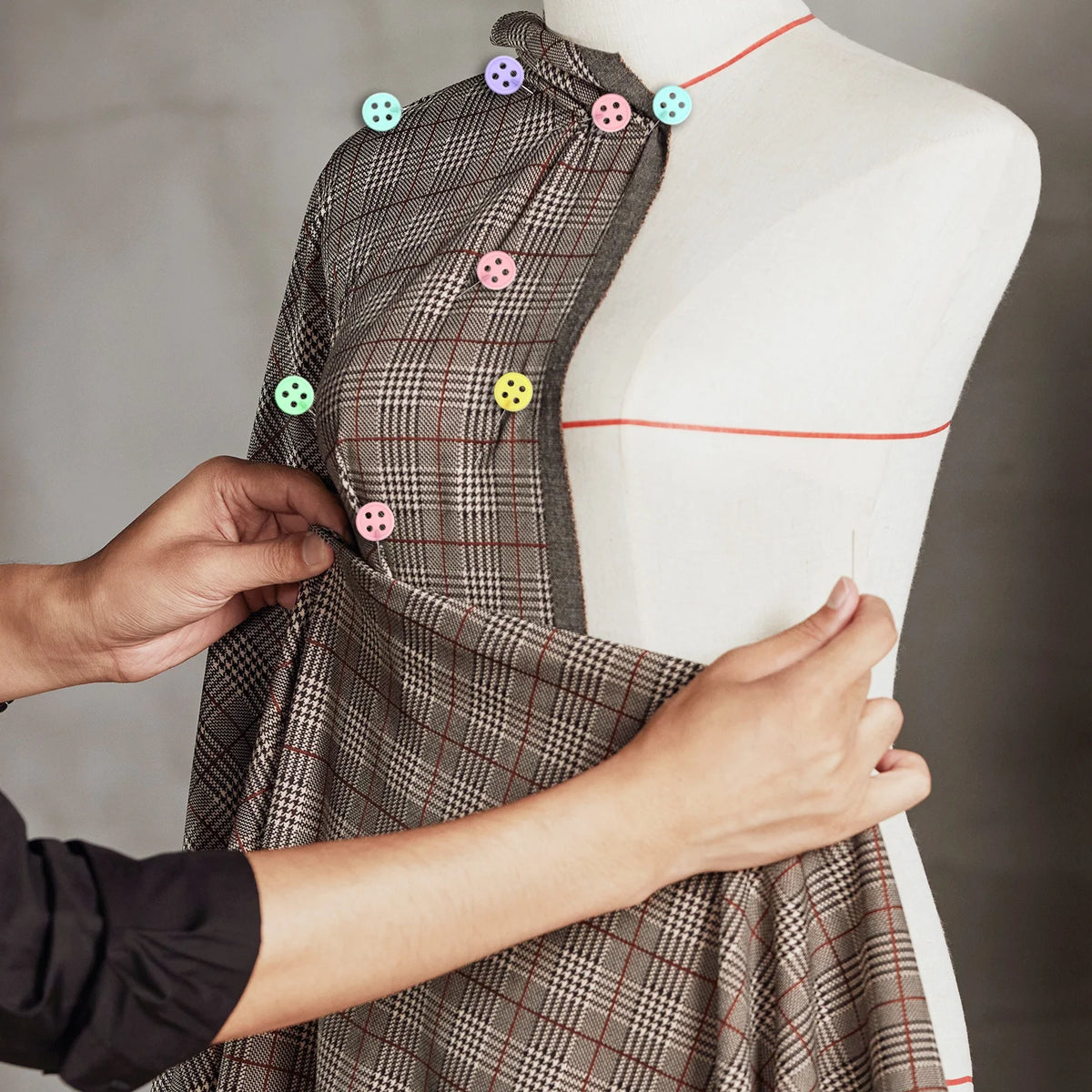 Hands are pinning a plaid dress onto a white mannequin using colorful nickel-plated dressmaking pins.