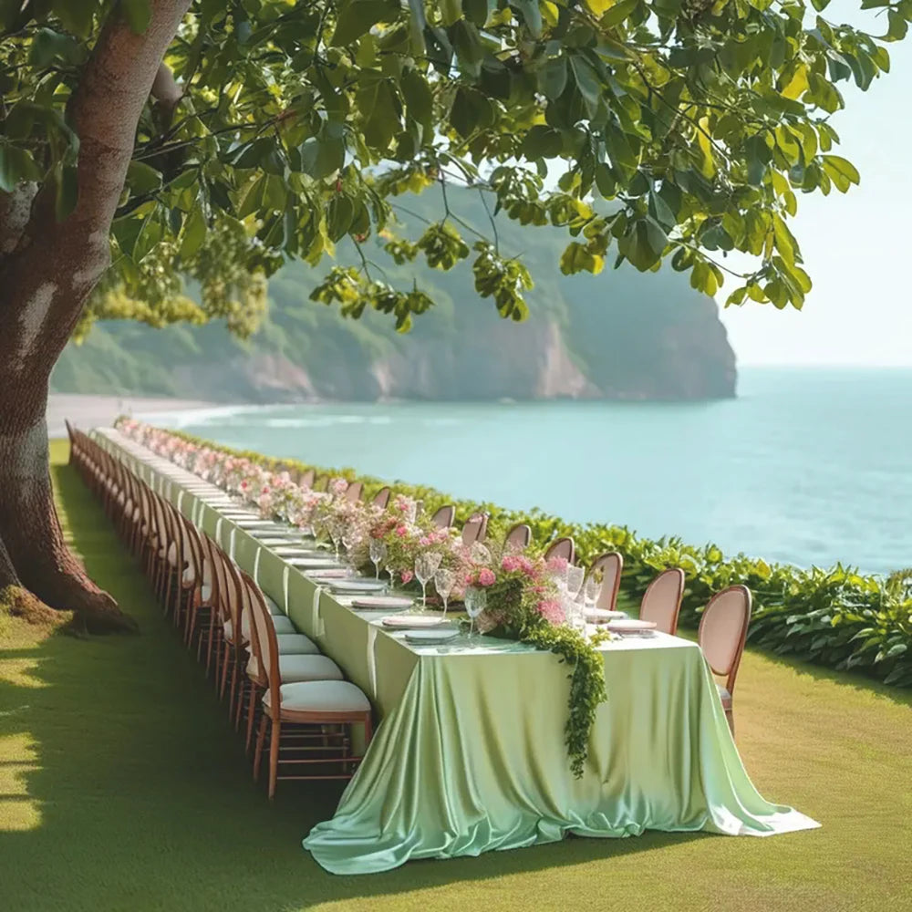 A long table set for an outdoor event with a satin green overlay cloth and pink floral arrangements overlooking the ocean.