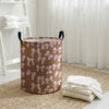 A brown canvas laundry basket with pink bow patterns sits on a beige rug next to white towels.