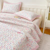 A floral quilted bedspread in pink and white patterns is displayed on a bed with a white headboard.