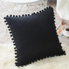 A black velvet decorative cushion cover with a black fringe resting on a white fluffy rug.
