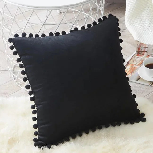 A black velvet decorative cushion cover with a black fringe resting on a white fluffy rug.