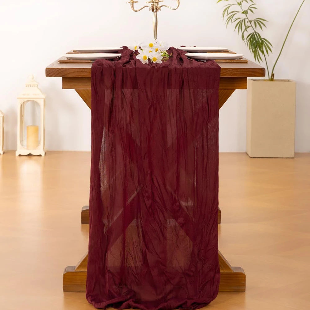 A deep burgundy cheesecloth table runner draped over a wooden table, decorated with white flowers.