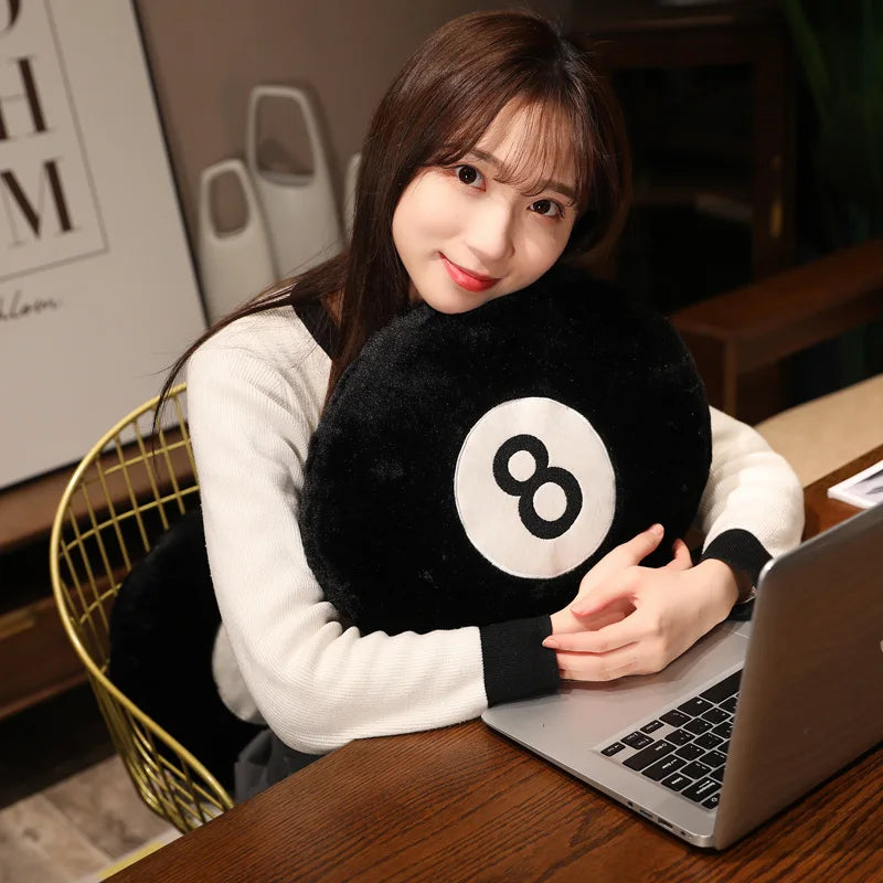 A woman is holding a black 8 ball plush pillow while working on a laptop.