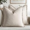 A close-up of a beige linen throw pillow with fringed edges resting on a light-colored sofa.