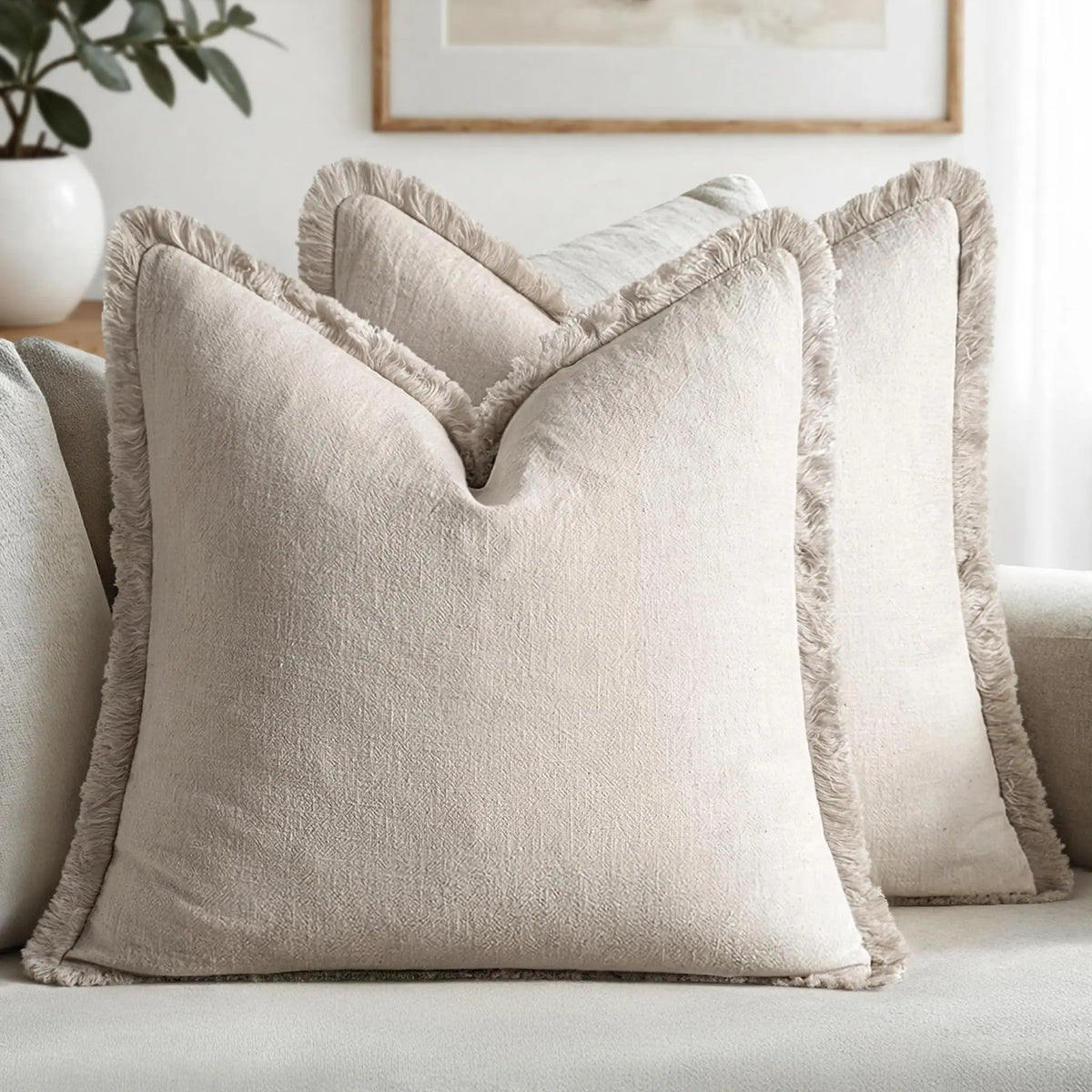 A close-up of a beige linen throw pillow with fringed edges resting on a light-colored sofa.