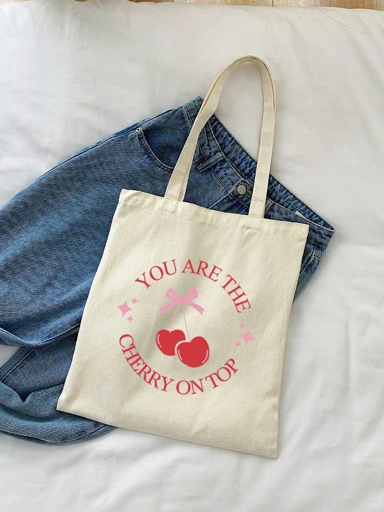 A beige canvas shopping bag with pink cherry embroidery on top, resting on a blue denim shirt.