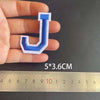 A hand holds a blue and white embroidered letter J iron-on patch next to a ruler showing its dimensions.