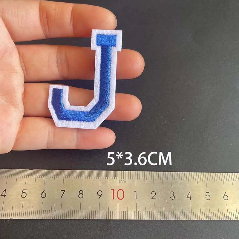 A hand holds a blue and white embroidered letter J iron-on patch next to a ruler showing its dimensions.
