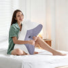 A woman is smiling while holding a white and purple ergonomic memory foam pillow on a white bed.