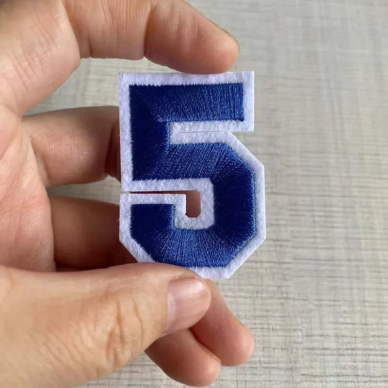 A hand holds a navy blue embroidered number 5 patch with white stitching.