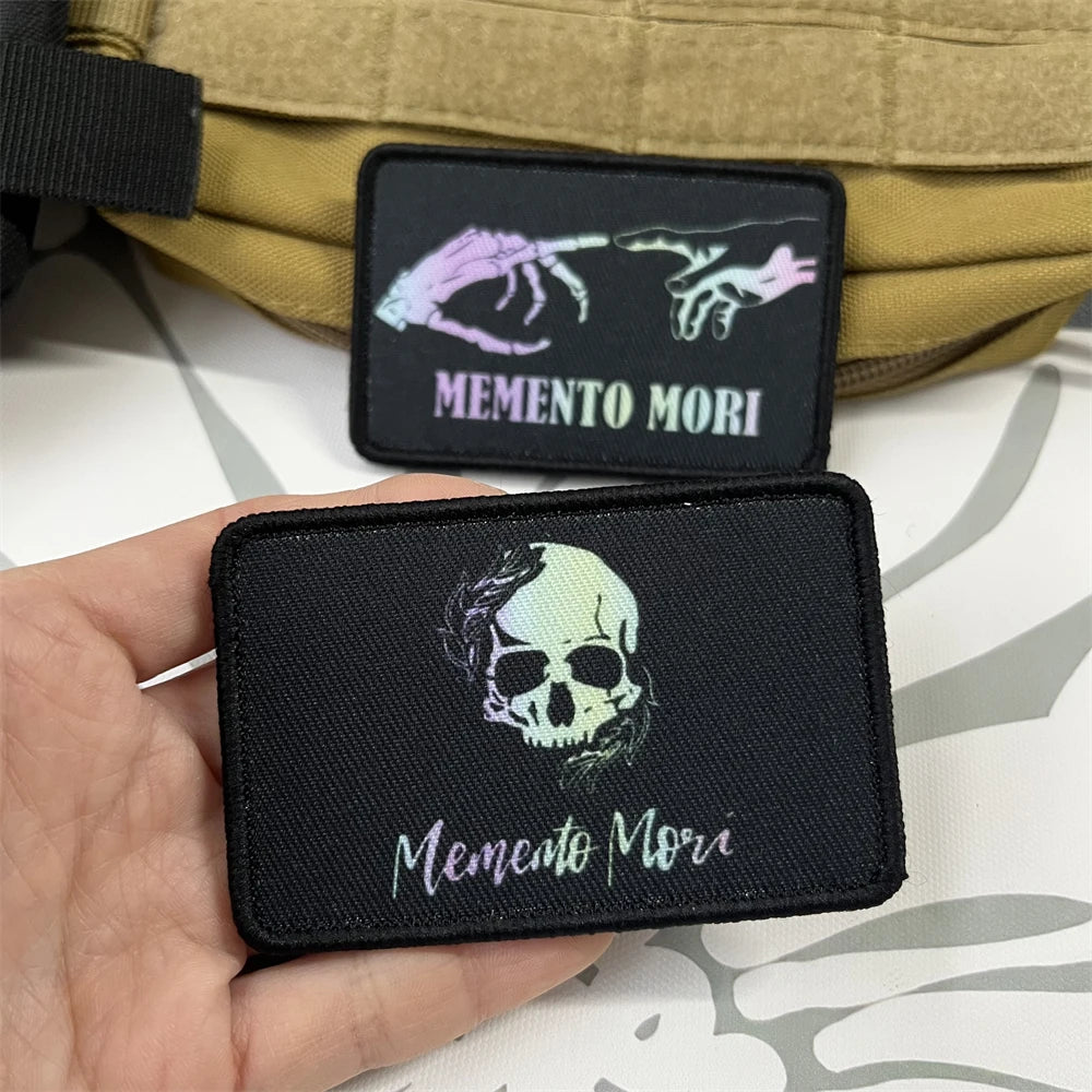 A hand holds a black badge patch featuring a skull and the text Memento Mori, with a tactical vest in the background.