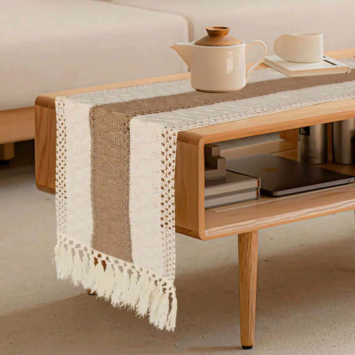 A beige linen cotton table runner with tassels is displayed on a wooden table next to a ceramic teapot.